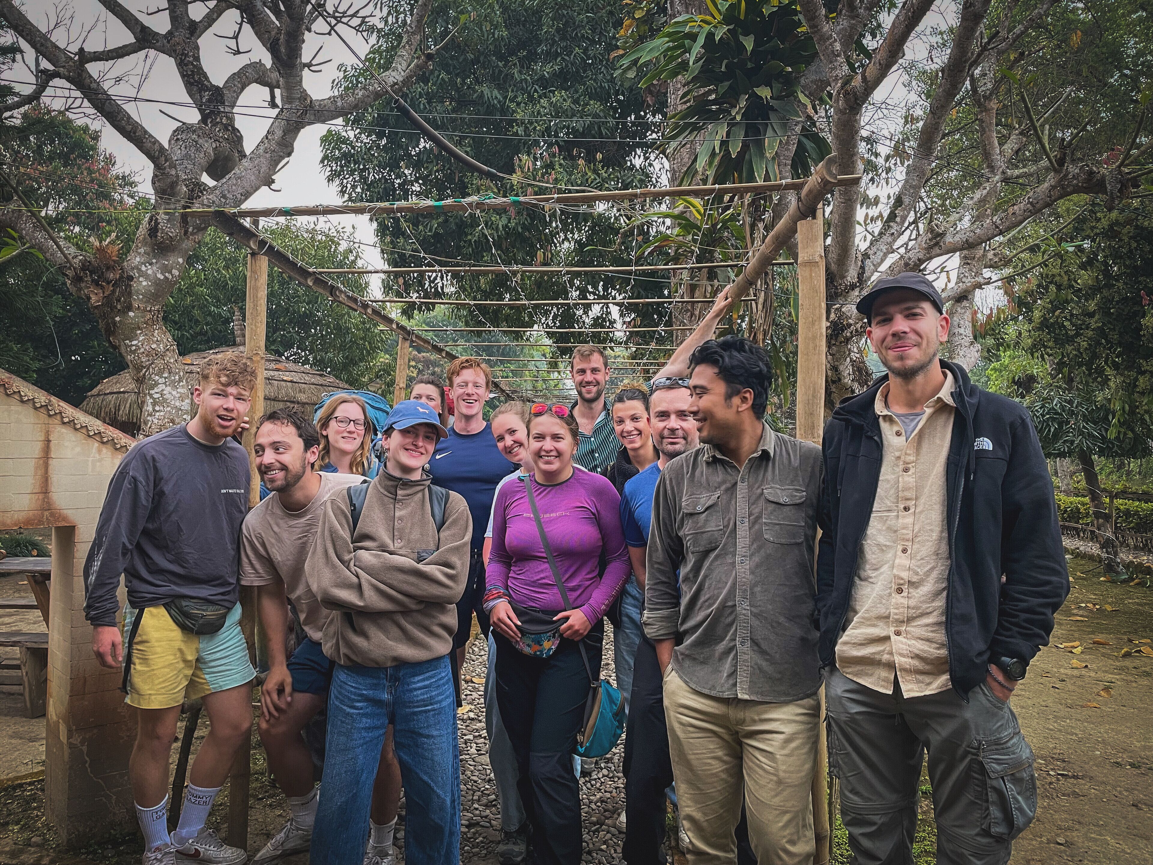 A group of people standing in an outdoor area with trees and a wooden structure overhead.