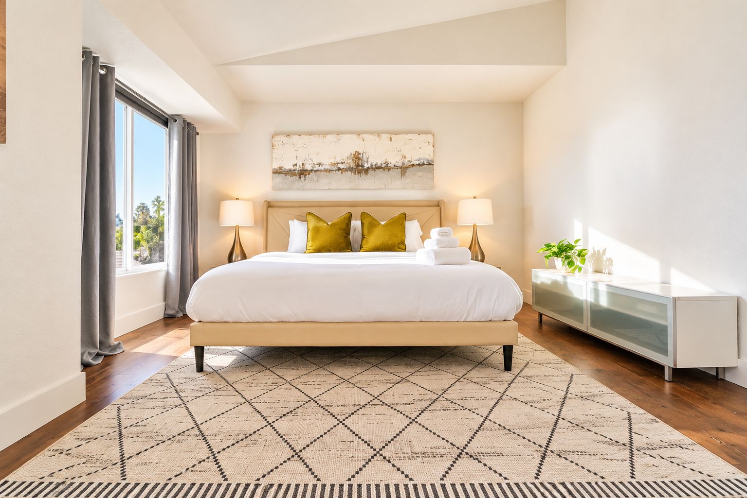 A modern hotel room with a large bed, a bedside table, a nightstand, a window with curtains, and a view of palm trees.