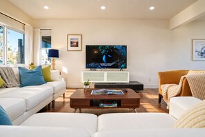 En moderne stue med et fladskærms-TV, hvide sofaer, et sofabord og en reol.