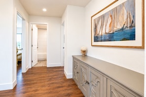 A hallway with wooden flooring, a white door leading to a bathroom, and a gray console with a painting of sailboats on the wall.