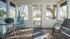 A covered porch with a table and chairs, a door leading outside, and a view of trees and a parking lot.
