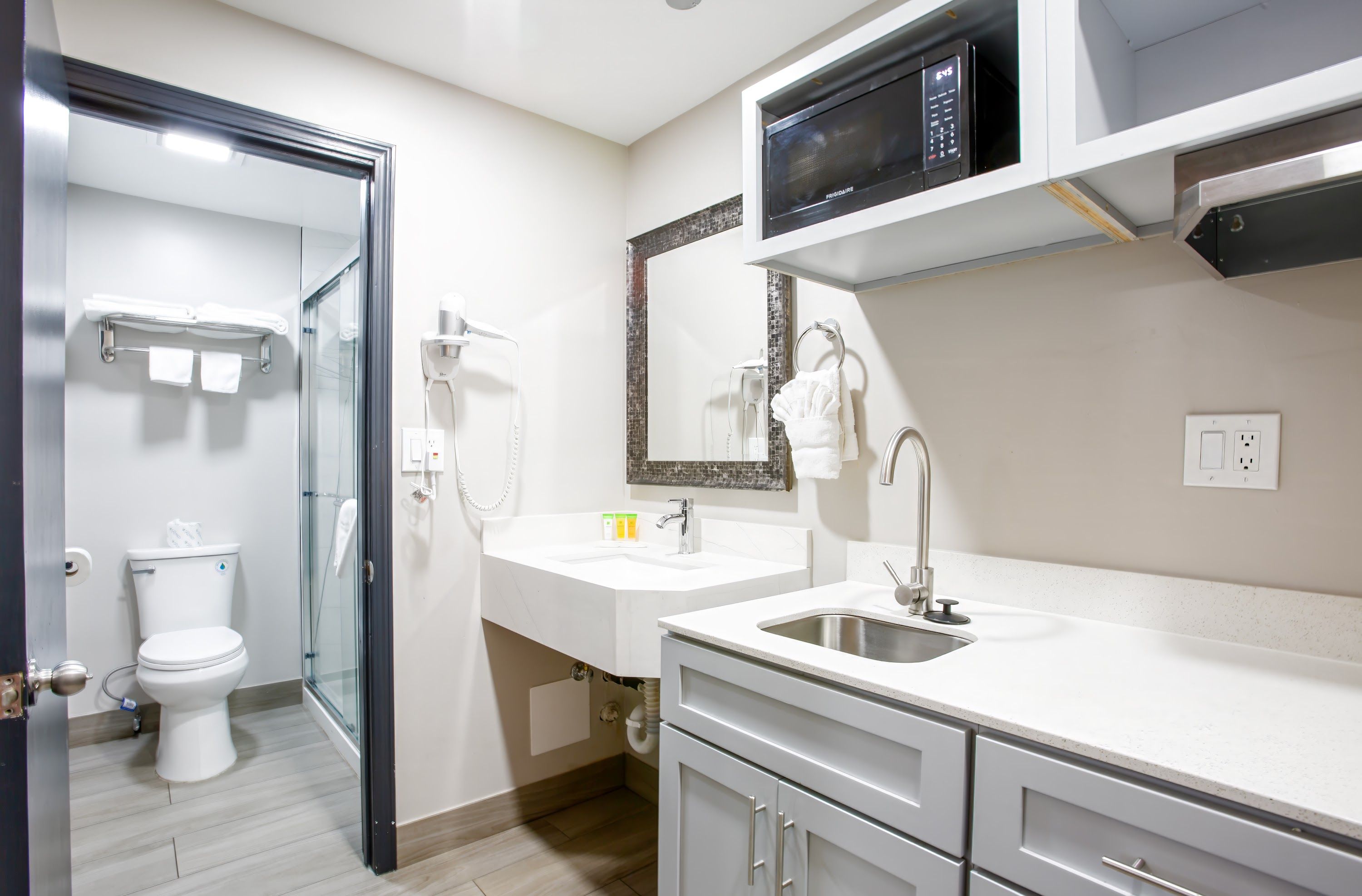 A modern bathroom with a toilet, a glass shower enclosure, a white sink with a mirror above, and a microwave above the sink.