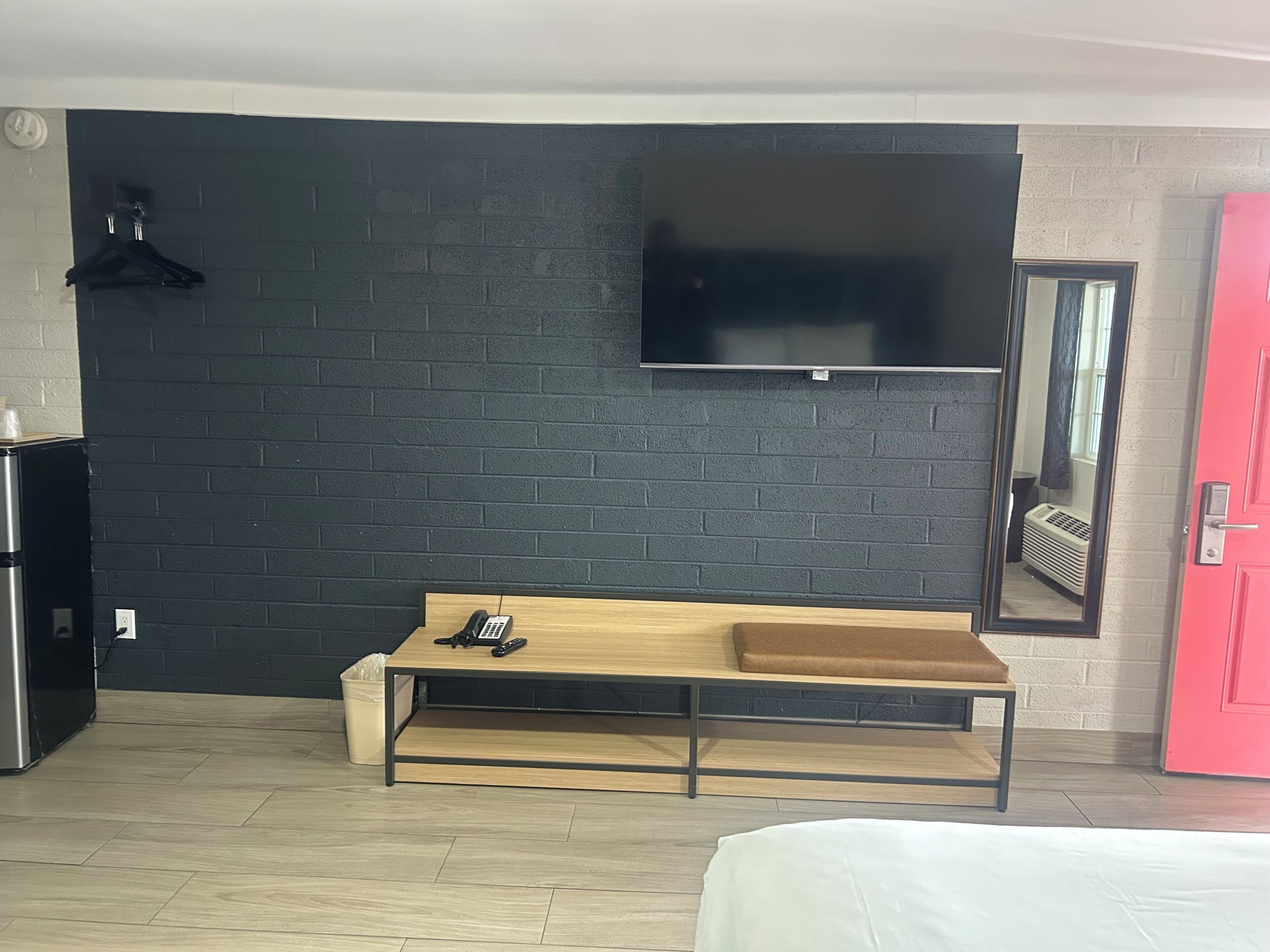 Une chambre moderne avec un mur en briques noires, un téléviseur fixé au mur, un banc en bois, un lit et une porte rouge.
