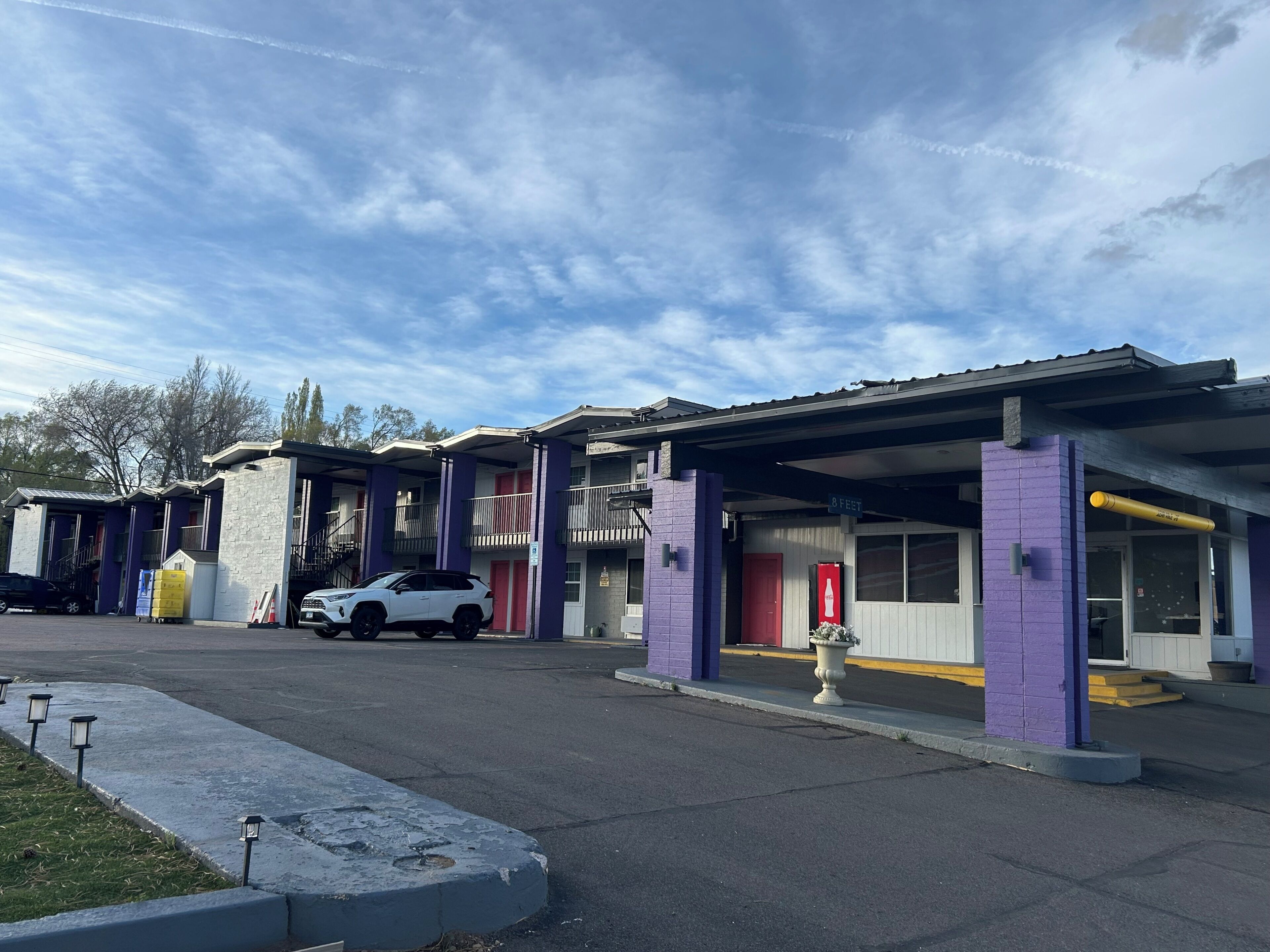 Motel amb una filera d'habitacions, una zona de pàrquing coberta i un SUV blanc aparcat al davant.