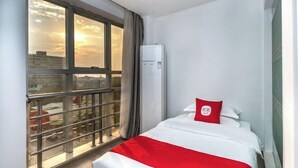 A hotel room with a bed, a window with a view of buildings, and an air conditioning unit. - Boutique Guodu Hotel (Nantong)