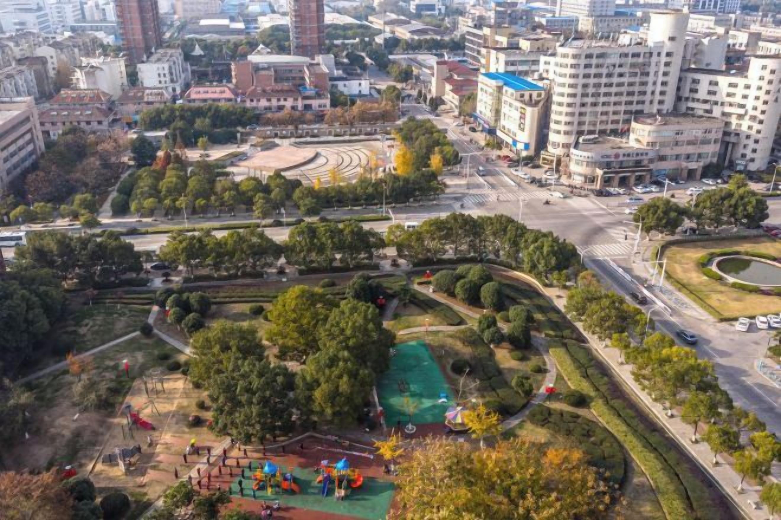 Vista aérea de un parque urbano con áreas de juegos, árboles y una carretera.