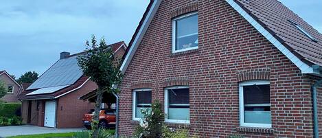 Une maison en briques rouges, avec un toit à pignon, un petit jardin planté de différentes espèces, et une allée pavée.