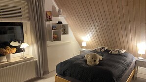 Un dormitorio acogedor con techo inclinado, una cama con colcha oscura, un oso de peluche en la cama, lámparas de noche y una ventana con persianas.