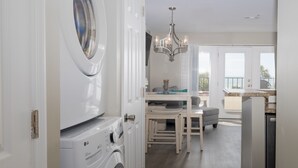 A laundry room with a front-loading washer and dryer, a dining area with a table and chairs, and a view of the outdoors through a large window. - Oceanfront Resort! at Ocean Delight! (North Topsail Beach)