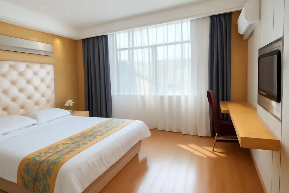 A hotel room with a bed, a TV, a desk, and a window with curtains.
