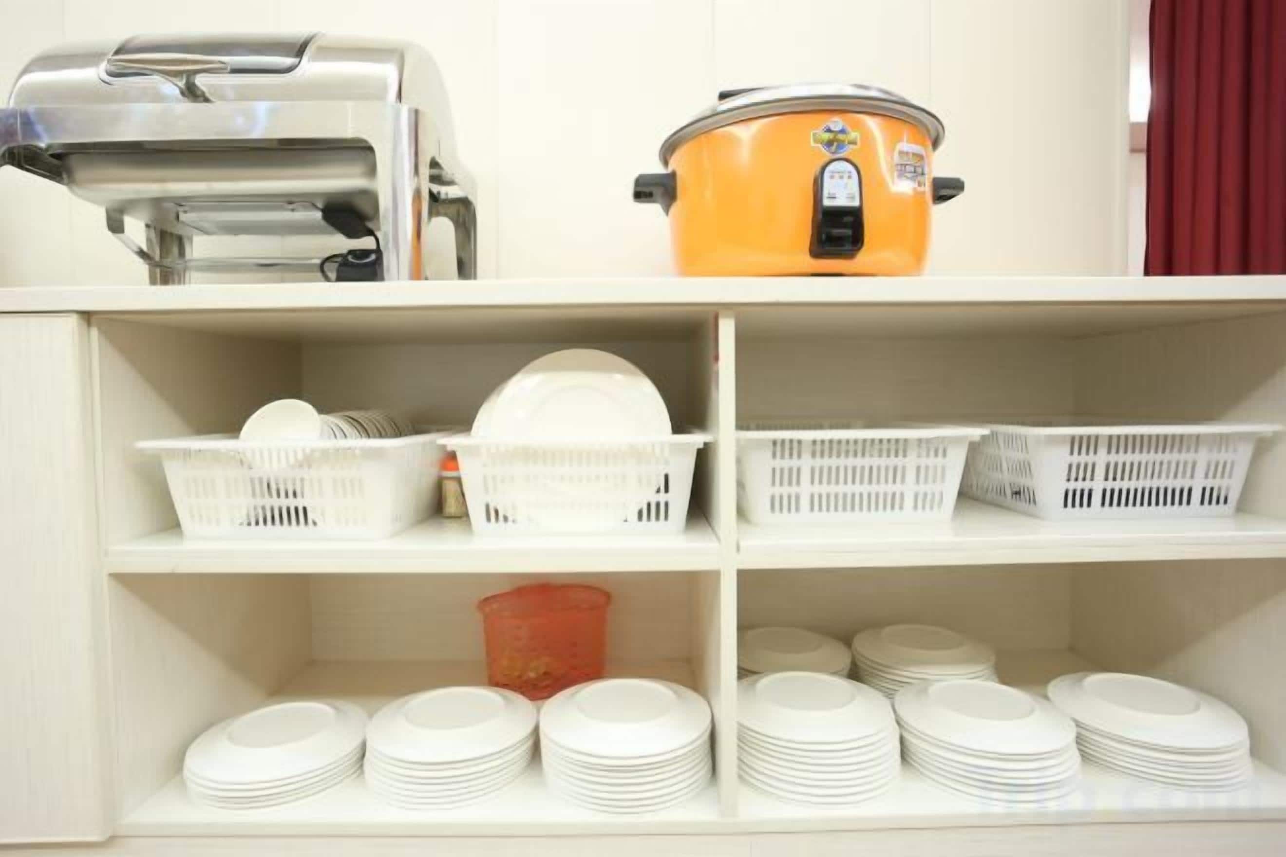 A kitchen area with a food processor, an orange slow cooker, and shelves with white plastic baskets and stacked plates.