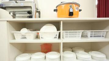 A kitchen area with a food processor, an orange slow cooker, and shelves with white plastic baskets and stacked plates.