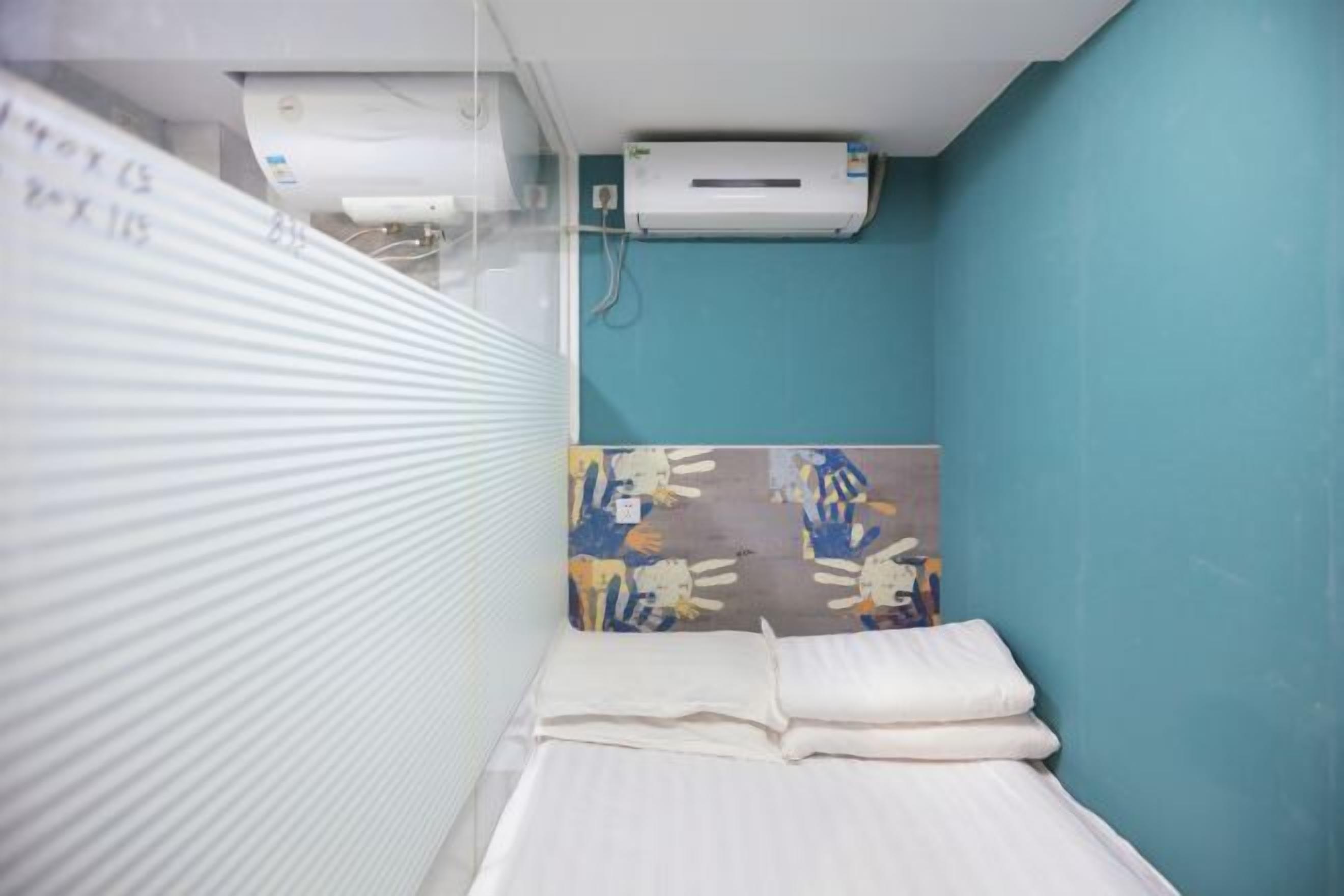 A small, private room with a bed, a window with blinds, an air conditioning unit, and a colorful artwork on the wall.