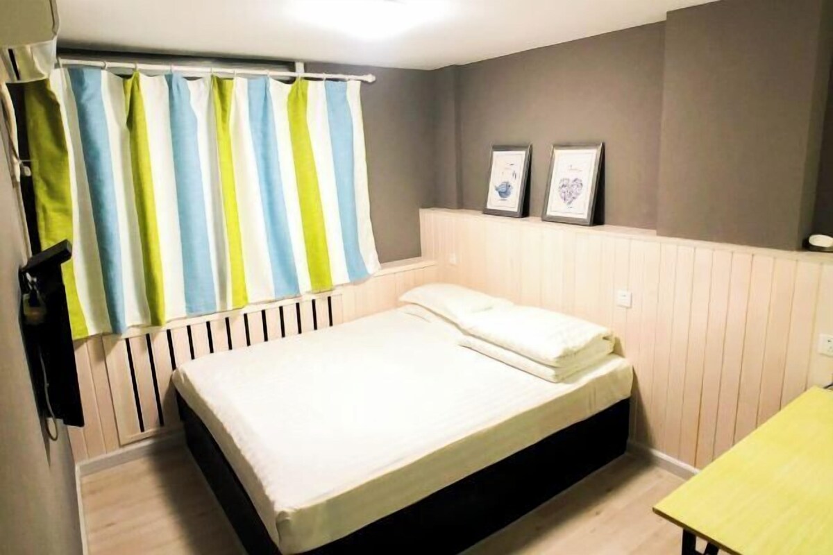 Un lit simple avec un matelas blanc, une tête de lit en bois, un rideau à rayures et deux tableaux encadrés accrochés au mur.