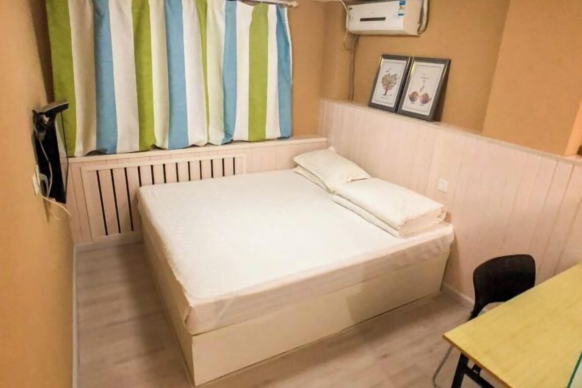 Una cama individual con colchón blanco, una mesita pequeña, una silla, un radiador y cortinas a rayas.