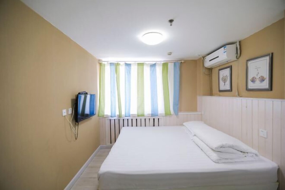 Une chambre d’hôtel avec un lit, une fenêtre avec des rideaux à rayures, un téléviseur fixé au mur et un radiateur.