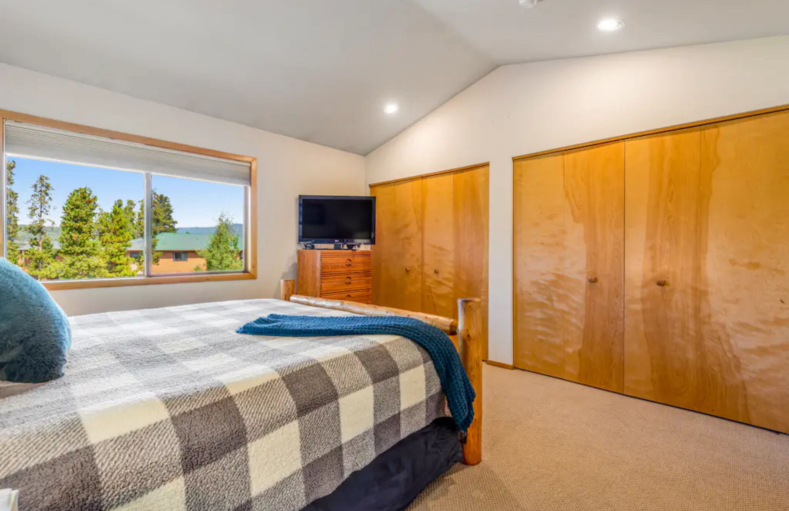 A bedroom with a large window, a bed with a checkered blanket, a TV on a wooden cabinet, and wooden wardrobes.