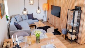 A modern living room with a wooden floor, grey sofa, and a dining area set for four. - Eggert Alm Apartments (Ilsenburg (Harz))