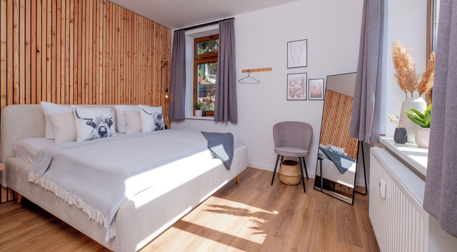 Un dormitorio moderno con pared de listones de madera, una cama grande con muchas almohadas, una mesita de noche, una silla y una ventana con cortinas.