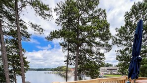 A wooden deck with two Adirondack chairs and a table, overlooking a lake with boats and a distant building. - Lakefront, Dock, Kayaks, Paddleboards, Pool,Hot tub and access to boat rentals! (Eatonton)
