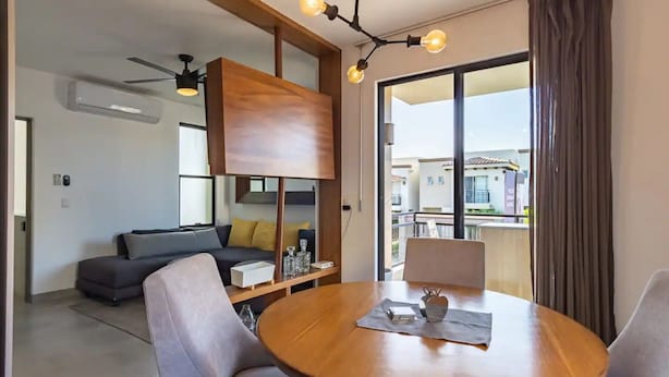 Un salón moderno con una mesa de comedor de madera, sofás grises y un ventanal con vistas a edificios.
