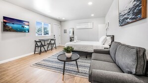 Ein modernes Wohnzimmer mit einem grauen Sofa, einem runden Couchtisch mit Pflanze, einem Streifen Teppich und einem Bett mit weißer Bettwäsche.
