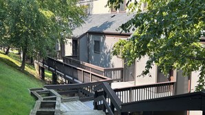 Eine hölzerne Treppe führt zu einem Gebäude mit einem grauen Dach, umgeben von Grünpflanzen und Bäumen.