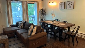 Ein Wohnzimmer mit einem braunen Ledersofa, einem Esstisch mit Stühlen, einem Blumentopf und einer Wandleuchte.