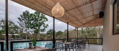 A covered outdoor dining area with tables and chairs, overlooking a pool and lush greenery.