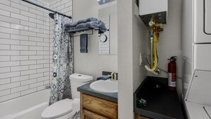 Une salle de bain avec des murs recouverts de carreaux blancs, des toilettes blanches, un lavabo avec un miroir, un porte-serviettes et un extincteur.