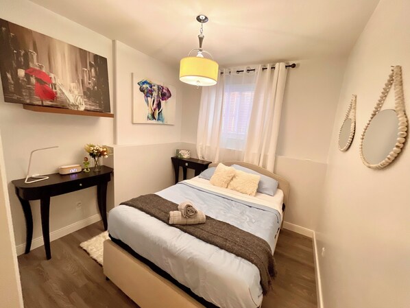 A bedroom with a bed, bedside tables, a desk, a lamp, a painting, and a window with curtains. - Luxury Suite-Unique Quotes Space (Saskatoon)