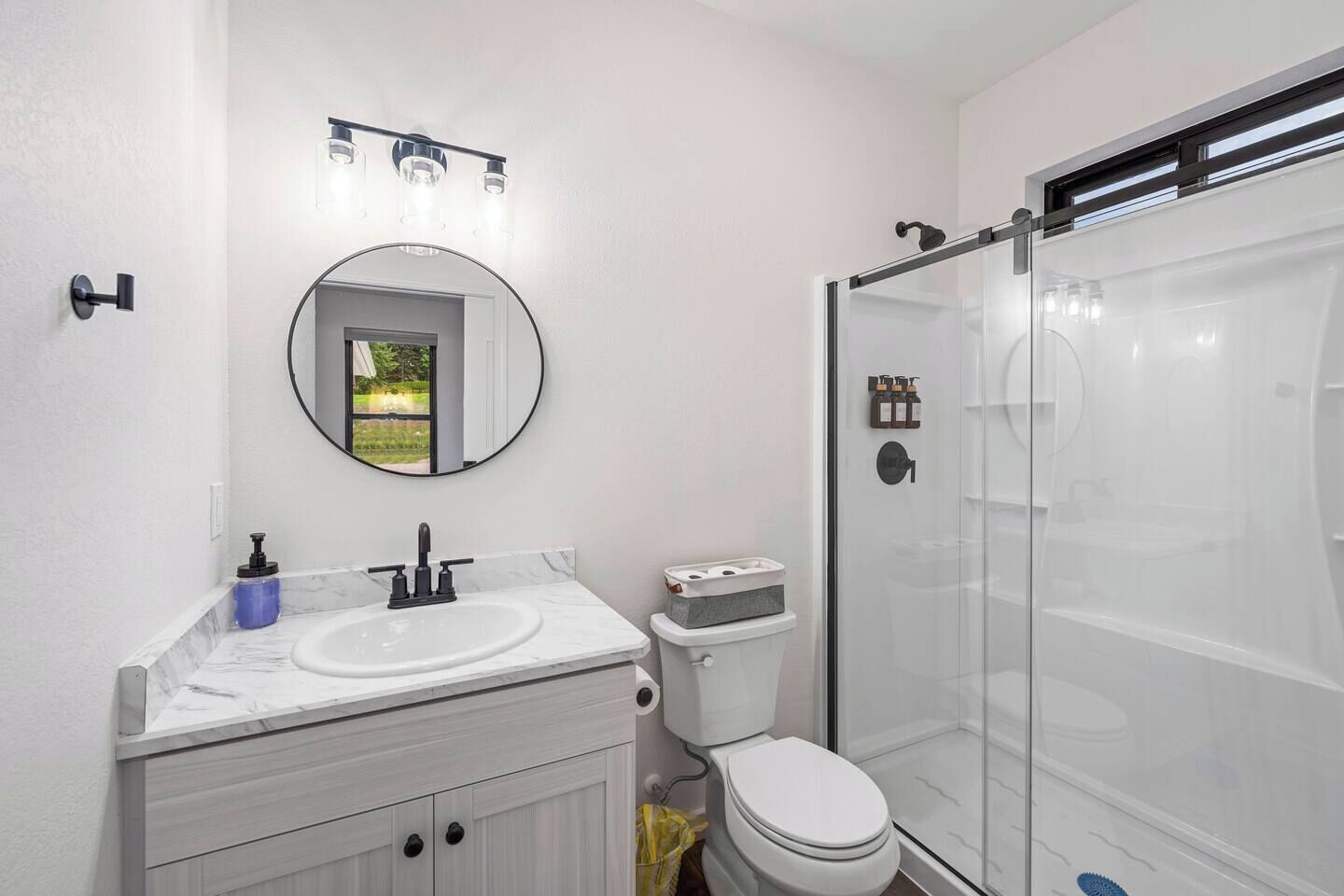 Une salle de bain avec un meuble-lavabo blanc, un miroir rond, des toilettes et une douche à parois de verre.