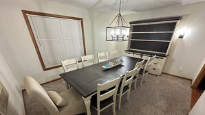 A dining area with a dark wooden table, white chairs, a chandelier, a window with blinds, and a built-in bench with storage.