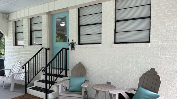 A porch with a teal door, a wooden bench, a small table, and a chair with teal cushions.