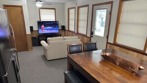 A living room with a wooden dining table, a beige sofa, and a television displaying a sports event.