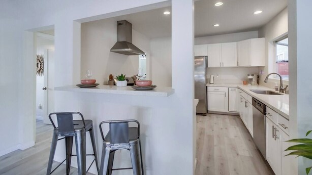 A modern kitchen with white cabinets, a stainless steel refrigerator, and a built-in oven. There are two bar stools and a small island with a range hood above. - Chic 3BR Spanish Bungalow in Walkable North Park (San Diego)