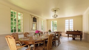 Un comedor con una gran mesa de madera dispuesta para seis personas, rodeado de sillas de mimbre, un piano de cola y ventanas amplias con vistas a la vegetación.