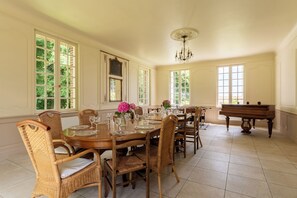 Ein Esszimmer mit einem großen Holztisch, gedeckt für sechs Personen, umgeben von Korbstühlen, einem Flügel und großen Fenstern mit Blick auf Grünflächen.