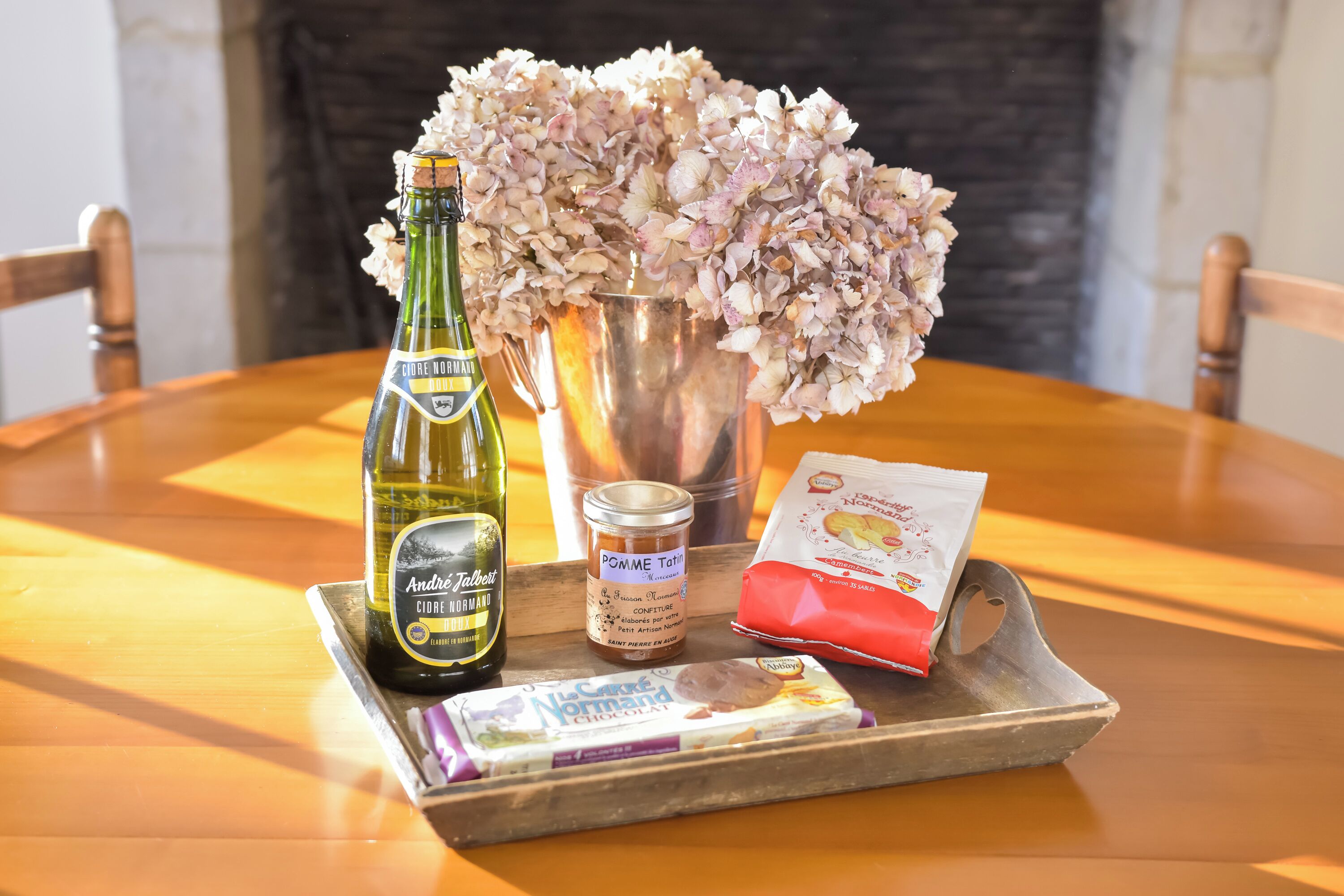 Un plateau avec une bouteille de cidre normand Andre Tallebert, un pot de confiture de pommes, un paquet de chocolat et un vase de fleurs, le tout posé sur une table en bois.