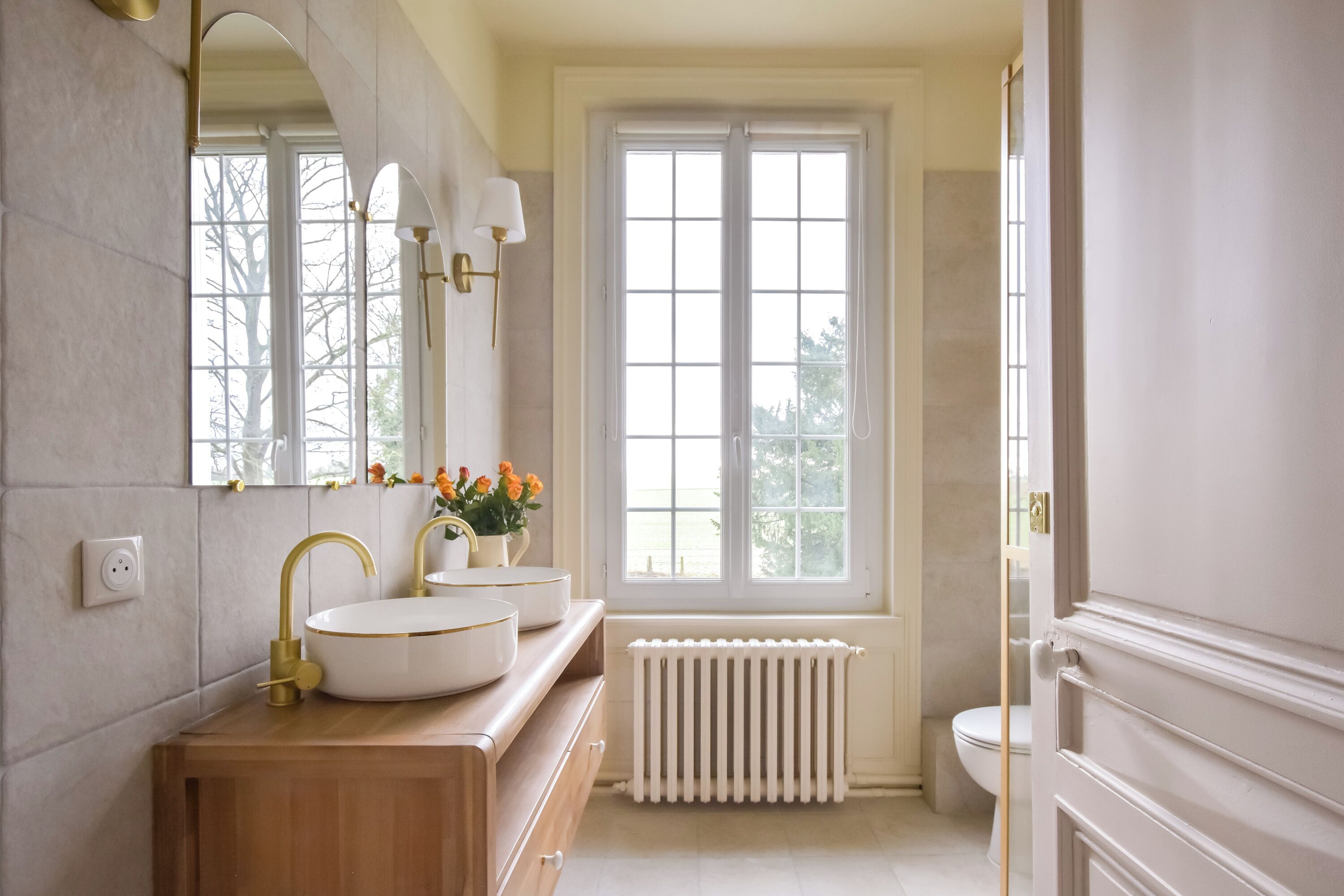 A bathroom with a large window, a white radiator, and a wooden vanity with a gold faucet.
