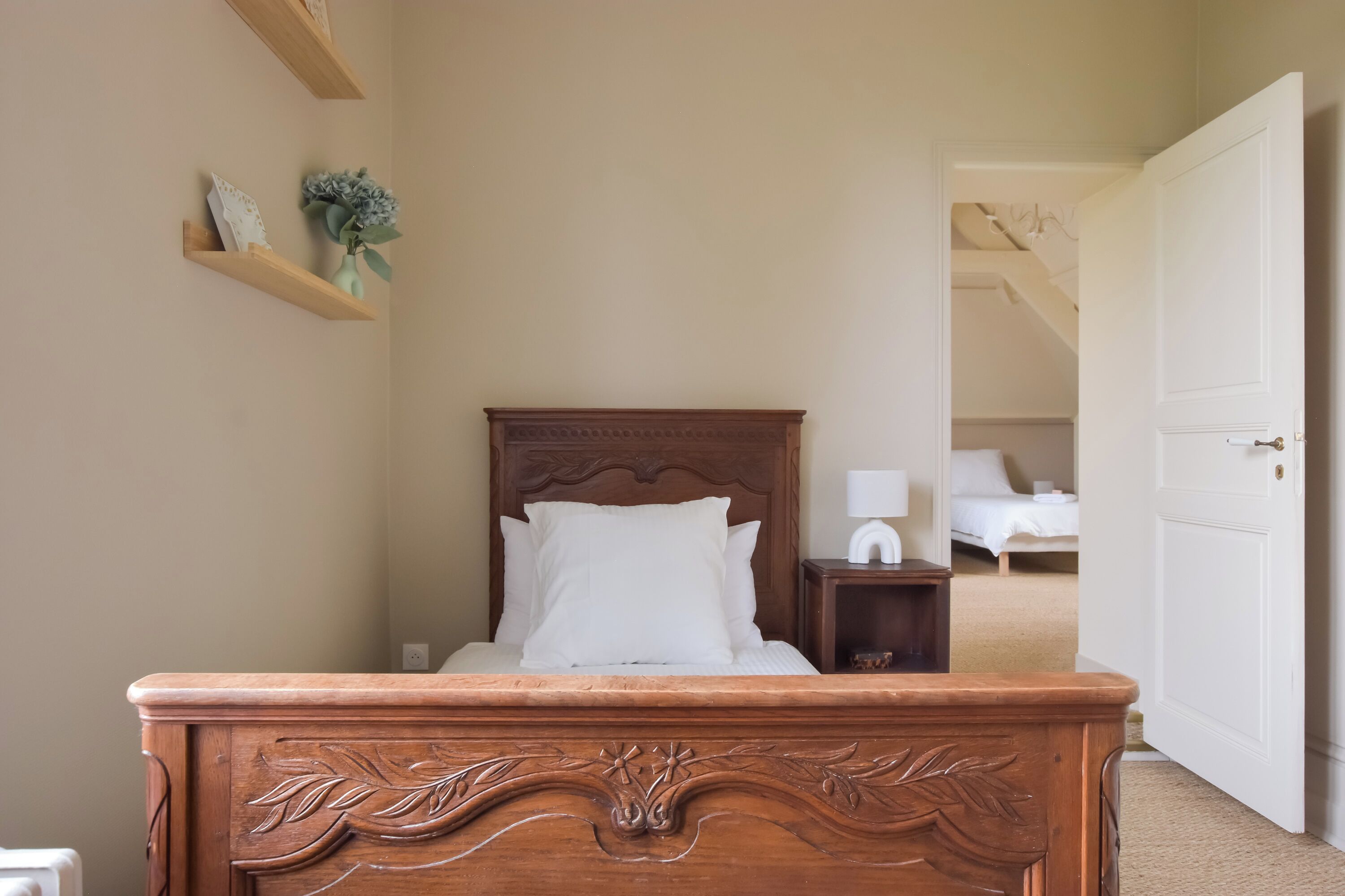 Una camera da letto con un letto di legno, un comodino, una porta bianca e un corridoio che porta verso un'altra stanza.