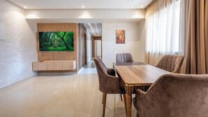 Un espace repas moderne avec une table en bois et des chaises rembourrées, un écran de télévision diffusant une scène de forêt et un tableau accroché au mur.