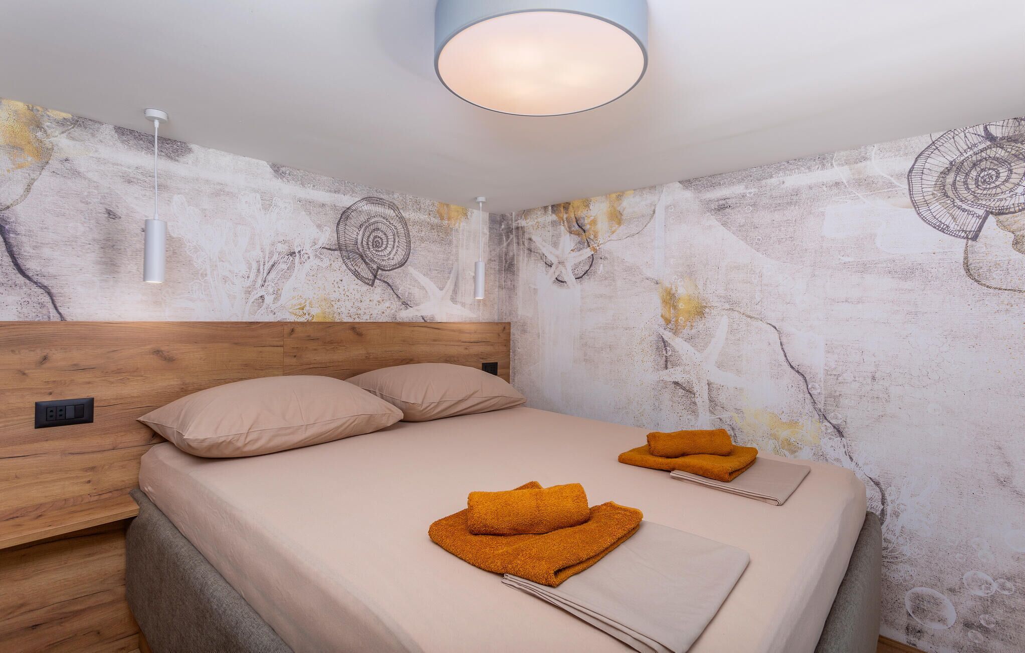 Et soveværelse med sengegavl i træ, to senge med beige sengetøj, et vægmaleri og en loftlampe.