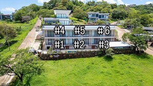 Un edificio moderno de varios pisos con unidades numeradas, rodeado de abundante vegetación y un cielo despejado.