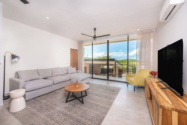 A modern living room with a grey sofa, a round coffee table, a yellow armchair, and a TV on a wooden stand.