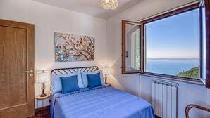 A bedroom with a bed, a nightstand, a lamp, a painting, and a window with a view of the sea.
