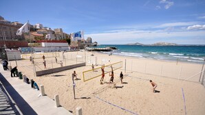 Una cancha de voleibol de playa con una red, rodeada de arena y un paisaje urbano al fondo.