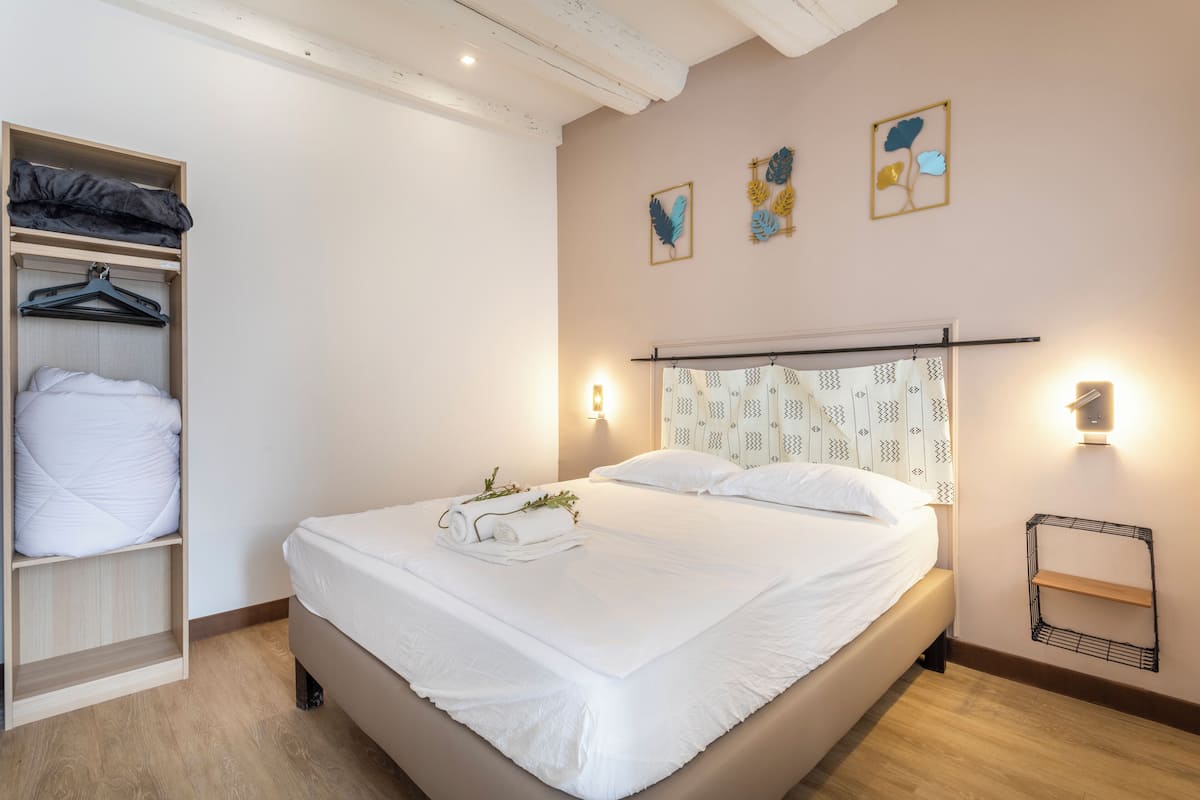 Un lit bien fait, avec une couette blanche et une serviette pliée, une armoire en bois où sont suspendus des vêtements, et une étagère fixée au mur.