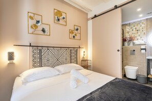 A hotel room with a bed, a headboard with a geometric pattern, wall art, a bathroom with a tiled shower, and a towel rack. - Adira Marseille Panier (Marseille)