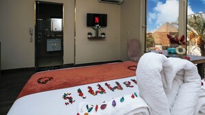 Une chambre d’hôtel avec un lit, un téléviseur fixé au mur, une fenêtre donnant sur une pyramide et une salle de bain visible à travers une porte ouverte.